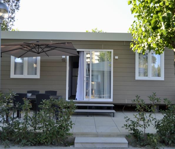 Modern mobile home with patio, table, and umbrella, surrounded by greenery.